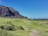 A lush green golf course is set against a backdrop of rugged cliffs under a clear blue sky. Several palm trees and shrubs dot the landscape, adding to the tropical atmosphere. The ocean can be seen in the distance, enhancing the scenic beauty.