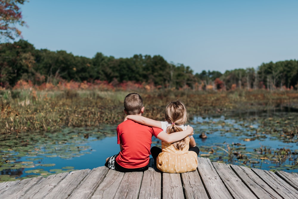a couple of kids sitting on a dock by a lake