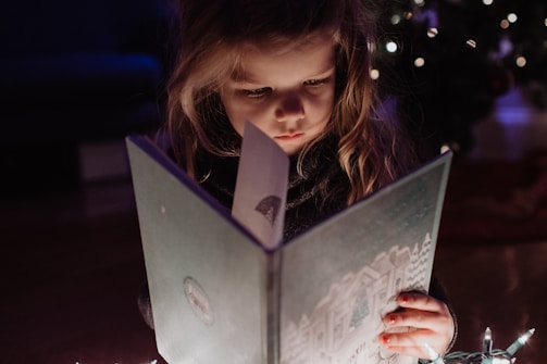A young child with long hair is intently reading a book. The scene is dimly lit, with warm light highlighting the child's focused expression. In the background, blurred lights suggest a festive or cozy setting, possibly a Christmas tree.