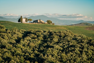 A picturesque rural landscape featuring a small chapel and a farmhouse surrounded by lush green fields. The distant rolling hills and a mountain range provide a serene backdrop beneath a clear blue sky with a few scattered clouds. Tall cypress trees stand near the buildings, adding to the tranquil environment.
