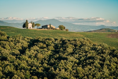 A picturesque rural landscape featuring a small chapel and a farmhouse surrounded by lush green fields. The distant rolling hills and a mountain range provide a serene backdrop beneath a clear blue sky with a few scattered clouds. Tall cypress trees stand near the buildings, adding to the tranquil environment.