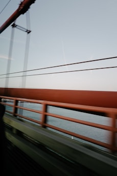 A bridge with sturdy metal beams is visible alongside railings, set against a backdrop of a clear sky and body of water. There are horizontal lines created by the bridge's structure, and the view appears to be taken from a moving vehicle.