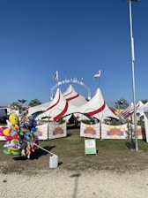 A large circus tent with red and white stripes is set up on a grassy area. Several flags fly atop the tent, and white chairs are visible inside. A collection of colorful helium balloons featuring various cartoon characters is positioned near the entrance. Banners with the logo 'Niuman Circus' are displayed at the front of the tent, and a tall streetlight stands on the right side of the image.