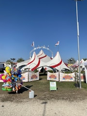 A large circus tent with red and white stripes is set up on a grassy area. Several flags fly atop the tent, and white chairs are visible inside. A collection of colorful helium balloons featuring various cartoon characters is positioned near the entrance. Banners with the logo 'Niuman Circus' are displayed at the front of the tent, and a tall streetlight stands on the right side of the image.