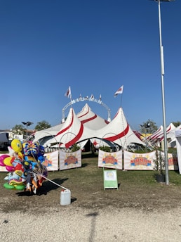 A large circus tent with red and white stripes is set up on a grassy area. Several flags fly atop the tent, and white chairs are visible inside. A collection of colorful helium balloons featuring various cartoon characters is positioned near the entrance. Banners with the logo 'Niuman Circus' are displayed at the front of the tent, and a tall streetlight stands on the right side of the image.