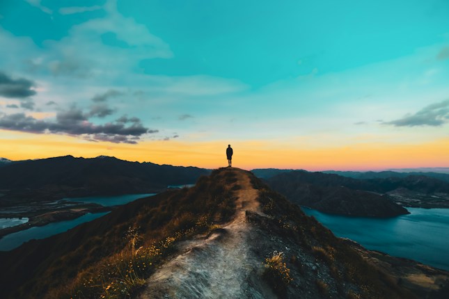 A solitary figure stands on a narrow mountain ridge at sunrise or sunset, overlooking a stunning panoramic landscape of hills, a lake, and a vast sky filled with clouds. The path is edged with grass and flowers, and the colors in the sky range from blue to orange and pink, casting a serene glow over the scene.
