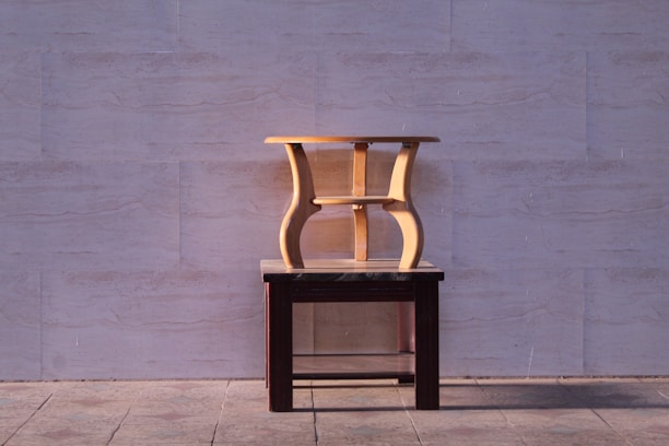 A wooden stool with a unique, twisting design is placed on top of a dark wooden table. The background consists of a light-colored stone wall, and the ground is made up of large stone tiles.