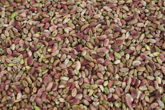 A close-up view of a large pile of shelled pistachio nuts, showcasing a variety of shades from green to brown and hints of reddish-purple.