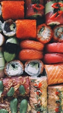 An assortment of sushi rolls arranged closely together, featuring a variety of ingredients including salmon, avocado, seaweed, and rice. Some rolls are topped with vibrant orange roe and garnished with herbs and slices of chili pepper.