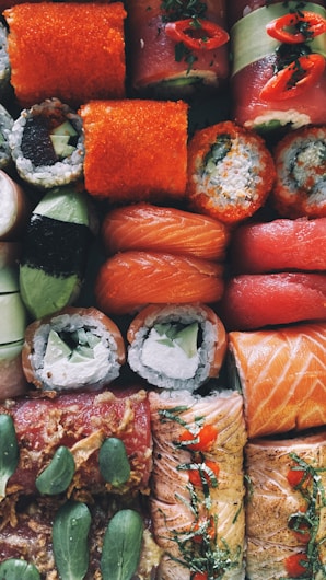 An assortment of sushi rolls arranged closely together, featuring a variety of ingredients including salmon, avocado, seaweed, and rice. Some rolls are topped with vibrant orange roe and garnished with herbs and slices of chili pepper.