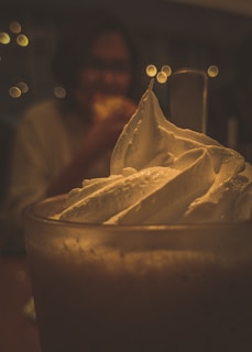 A close-up view of a glass filled with a creamy beverage topped with whipped cream. In the background, there is a blurred person who appears to be holding or eating something. The scene is softly lit with golden tones and out-of-focus lights that create a bokeh effect.