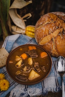 A bowl of hearty stew with chunks of meat, potatoes, and carrots is placed on a blue and white plaid fabric. Accompanying the stew is a rustic loaf of bread, a spoon, and a piece of corn on the cob, partially visible in the background. There are also yellow flowers near the arrangement, adding a touch of color.