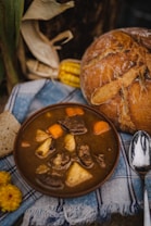 A bowl of hearty stew with chunks of meat, potatoes, and carrots is placed on a blue and white plaid fabric. Accompanying the stew is a rustic loaf of bread, a spoon, and a piece of corn on the cob, partially visible in the background. There are also yellow flowers near the arrangement, adding a touch of color.