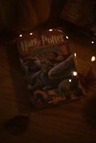A book titled 'Harry Potter and the Prisoner of Azkaban' lies on a wooden surface. The cover features a depiction of a mythical creature along with a person riding it. The setting is dimly lit with small, warm-colored fairy lights adding a cozy ambiance. Autumn leaves are scattered around, enhancing a seasonal atmosphere.