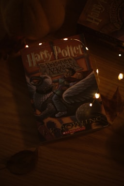 A book titled 'Harry Potter and the Prisoner of Azkaban' lies on a wooden surface. The cover features a depiction of a mythical creature along with a person riding it. The setting is dimly lit with small, warm-colored fairy lights adding a cozy ambiance. Autumn leaves are scattered around, enhancing a seasonal atmosphere.