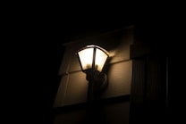 A wall-mounted outdoor lantern emits a warm glow, illuminating the wooden paneling of a building exterior. The lighting creates soft shadows on the textured wall surface, accentuating its grooves, while the rest of the surroundings remain dark.