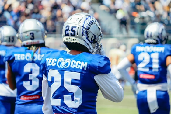 Several American football players are on a field, wearing blue jerseys with white numbers and silver helmets. The focal point is a player with the number 25 on their back. Other players are nearby, also in similar gear, with indistinct people in the background suggesting a stadium environment.