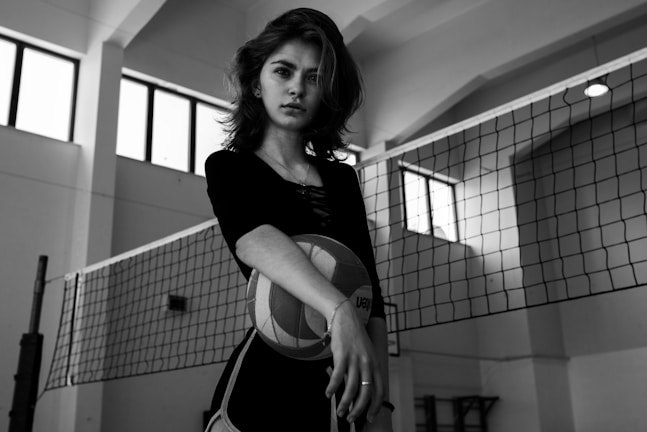 A person stands in what appears to be an indoor volleyball court, holding a volleyball. The environment is well-lit with natural light coming through large windows, creating a dramatic contrast in the black-and-white image.