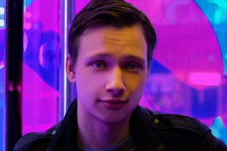 A young person with short hair stands in front of a vibrant neon-lit background. The colors pink and blue dominate the backdrop, creating a colorful and modern atmosphere. The individual is wearing a dark jacket and looks directly at the camera with a neutral expression.