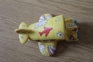 A yellow toy airplane with a whimsical design featuring stars, moons, clouds, and sun illustrations. The airplane is covered with various colorful images like red airplanes and blue clouds. It has a blue propeller at the front and is placed on a wooden surface.
