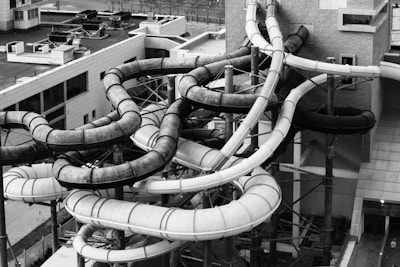 A complex network of twisting and turning water slides attached to a building. The slides are made of tubes, some dark and some light in color, and are supported by metal scaffolding. The environment appears industrial with the surrounding area consisting of other buildings and urban structures.