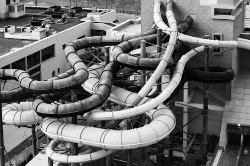 A complex network of twisting and turning water slides attached to a building. The slides are made of tubes, some dark and some light in color, and are supported by metal scaffolding. The environment appears industrial with the surrounding area consisting of other buildings and urban structures.