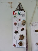 A vertical poster features a collection of small rock samples attached to a white background. Each rock is outlined with colorful glitter, and labels such as 'Marble' and 'Schist' identify them. The title 'Rocks World House' is handwritten at the top in multicolored letters. The poster is suspended by a braided cord.