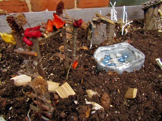 A miniature garden setting with a makeshift structure made from twigs and decorated with vibrant dried flowers. A small, rustic pond is crafted using a shallow dish partially filled with decorative stones. The garden is surrounded by soil and bark, giving a natural and earthy appearance. In the background, there's a log cabin model and small tree branches painted white.