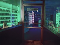A dimly lit storefront with a glass door displaying a 'Closed' sign. Shelves lined with electronic devices can be seen inside, illuminated by ambient green and blue lighting. Various gadgets and boxes are arranged on the display shelves.