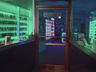 A dimly lit storefront with a glass door displaying a 'Closed' sign. Shelves lined with electronic devices can be seen inside, illuminated by ambient green and blue lighting. Various gadgets and boxes are arranged on the display shelves.