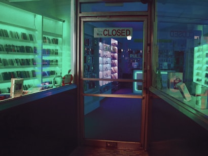 A dimly lit storefront with a glass door displaying a 'Closed' sign. Shelves lined with electronic devices can be seen inside, illuminated by ambient green and blue lighting. Various gadgets and boxes are arranged on the display shelves.