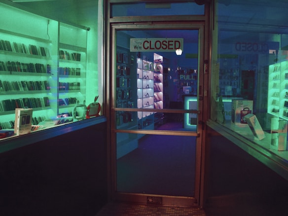 A dimly lit storefront with a glass door displaying a 'Closed' sign. Shelves lined with electronic devices can be seen inside, illuminated by ambient green and blue lighting. Various gadgets and boxes are arranged on the display shelves.