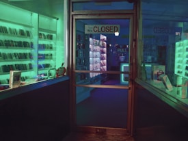 A dimly lit storefront with a glass door displaying a 'Closed' sign. Shelves lined with electronic devices can be seen inside, illuminated by ambient green and blue lighting. Various gadgets and boxes are arranged on the display shelves.