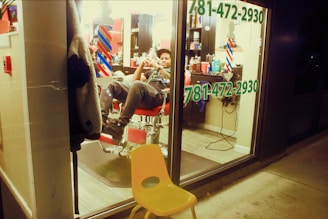 A person in a relaxed pose sits inside a barbershop, visible through a large window. The interior is lit with warm lights, and typical barbershop elements like barber poles and various hair products on shelves can be seen. The window prominently displays a phone number in green text. Outside the shop, a yellow plastic chair is placed on the sidewalk.