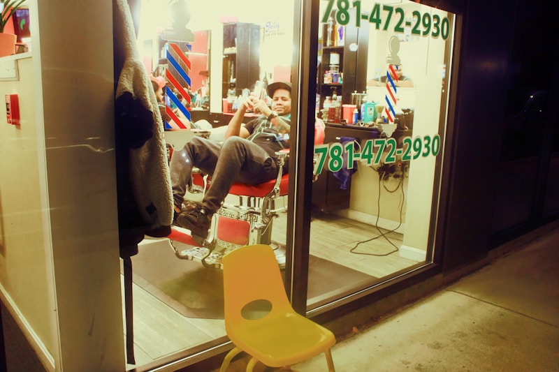 A person in a relaxed pose sits inside a barbershop, visible through a large window. The interior is lit with warm lights, and typical barbershop elements like barber poles and various hair products on shelves can be seen. The window prominently displays a phone number in green text. Outside the shop, a yellow plastic chair is placed on the sidewalk.
