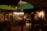 Outdoor restaurant seating area at night, with dim warm lighting and a cozy atmosphere. Surroundings include plants, decorative lighting, and an illuminated sign. A few people are visible in the background, suggesting a lively, yet intimate setting.