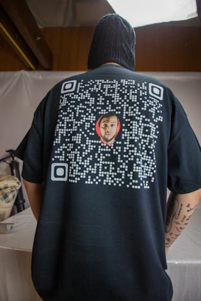 A person is seen from the back wearing a black shirt featuring a QR code design with a small image of a man's face in the center. The person is indoors, with a neutral colored backdrop and appears to have tattoos on their arm.