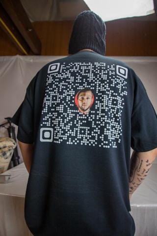 A person is seen from the back wearing a black shirt featuring a QR code design with a small image of a man's face in the center. The person is indoors, with a neutral colored backdrop and appears to have tattoos on their arm.