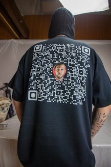 A person is seen from the back wearing a black shirt featuring a QR code design with a small image of a man's face in the center. The person is indoors, with a neutral colored backdrop and appears to have tattoos on their arm.