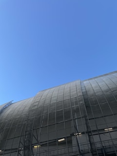 A large, curved industrial or commercial building structure with a glass or metal grid facade against a clear blue sky. The structure features a grid pattern and appears modern and industrial.