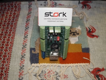 A green and blue circuit board module with various electronic components is placed on a multicolored woven fabric. The circuit board is labeled 'stork' and has various switches and connectors. The background fabric has intricate patterns in red, beige, and other colors.