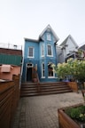 A charming, two-story blue building with white trim features a steep gabled roof. Wooden steps lead to the entrance, surrounded by a neatly fenced area and potted plants. The setting includes adjacent buildings and a small garden area with flowering shrubs.