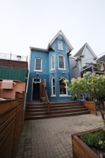 A charming, two-story blue building with white trim features a steep gabled roof. Wooden steps lead to the entrance, surrounded by a neatly fenced area and potted plants. The setting includes adjacent buildings and a small garden area with flowering shrubs.