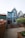 A charming, two-story blue building with white trim features a steep gabled roof. Wooden steps lead to the entrance, surrounded by a neatly fenced area and potted plants. The setting includes adjacent buildings and a small garden area with flowering shrubs.