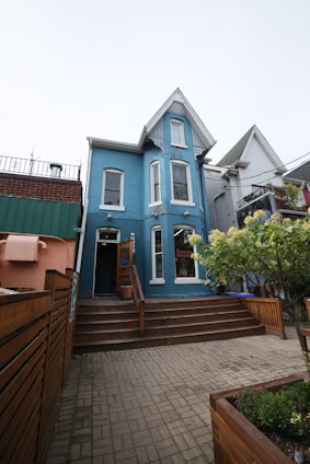 A charming, two-story blue building with white trim features a steep gabled roof. Wooden steps lead to the entrance, surrounded by a neatly fenced area and potted plants. The setting includes adjacent buildings and a small garden area with flowering shrubs.