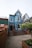 A charming, two-story blue building with white trim features a steep gabled roof. Wooden steps lead to the entrance, surrounded by a neatly fenced area and potted plants. The setting includes adjacent buildings and a small garden area with flowering shrubs.