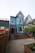 A charming, two-story blue building with white trim features a steep gabled roof. Wooden steps lead to the entrance, surrounded by a neatly fenced area and potted plants. The setting includes adjacent buildings and a small garden area with flowering shrubs.