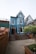 A charming, two-story blue building with white trim features a steep gabled roof. Wooden steps lead to the entrance, surrounded by a neatly fenced area and potted plants. The setting includes adjacent buildings and a small garden area with flowering shrubs.