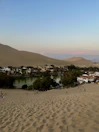 A serene desert oasis surrounded by sand dunes with a body of water in the center, bordered by lush greenery and small buildings. People are visible walking on the sand dunes, and the sky above is a gradient of blue to soft pink.