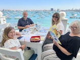 A family of four is seated at an outdoor restaurant table overlooking a bay filled with numerous boats. The group consists of two adults and two children. The sky is clear, and the city skyline is visible in the background.